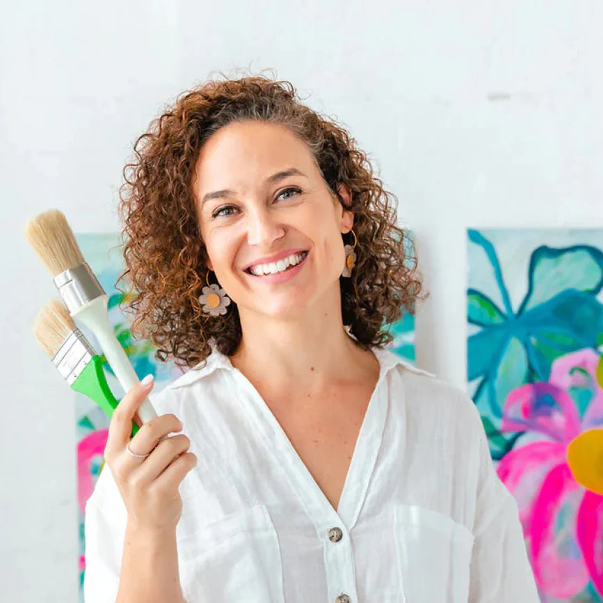 Woman holding paintbrushes with colorful floral artwork in the background